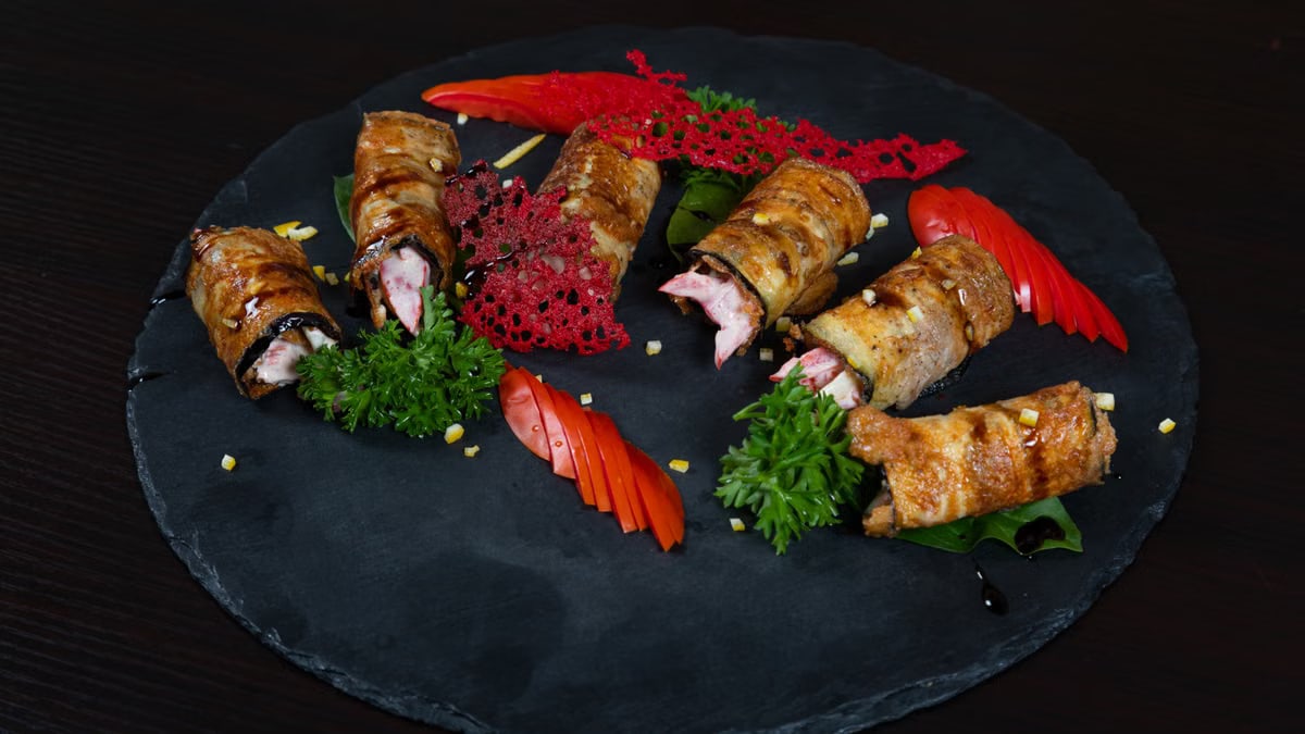 A gourmet dish of grilled meat rolls and fresh vegetables on a slate platter.